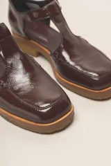 Zapato tipo guillermina de cuero charol color marrón oscuro, con tira T y hebilla ajustable en el lateral. Posee puntera cuadrada y suela de goma color caramelo con taco bajo.