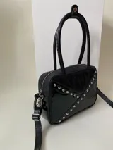 Cartera rígida negra con detalles en rojo y tachas plateadas. Tiene herrajes y cierre metálico plateado marca YKK, asa fina para llevar al hombro o en la mano y correa larga extra regulable. El interior es forrado, tiene un bolsillo con cierre enmarcado en cuero, y un segundo bolsillo plaqué.