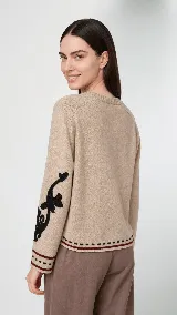 Sweater de lana color beige con calce holgado y cuello redondo. Presenta un bordado negro con diseño de arabescos en una de las mangas y detalles de rayas y puntos en contraste en el ruedo y los puños.