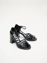 Sandalias de taco alto ancho con diseño retro, confeccionadas en charol negro. Presentan tiras delanteras múltiples entrelazadas, talón cerrado y una correa fina ajustable al tobillo con hebilla metálica.
