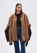Poncho de tejido grueso color camel, con bordes y mangas en color marrón oscuro. Cierre frontal con broche rectangular negro.