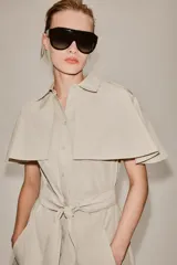 Vestido camisero midi de algodón con diseño de capa, cuello solapa, manga corta tipo capa y trabillas en los hombros. Presenta cierre frontal con botones, bolsillos delanteros y cinturón ajustable del mismo tejido con lazada. El bajo tiene un acabado en línea evasé.