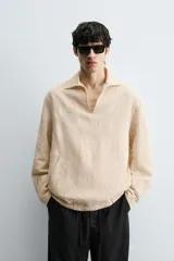 Camisa de manga larga color beige claro con textura jacquard arrugada, cuello solapa con abertura en V y bolsillo canguro frontal. Posee cordones ajustables en el bajo lateral.