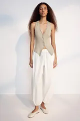 Top de algodón color beige con escote en V, sin mangas y cierre frontal con botones. Presenta un diseño asimétrico en el bajo con abertura delantera.