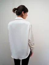 Camisa de manga larga confeccionada en satén de color blanco, con superficie lisa, leve brillo y caída suave. Presenta cuello clásico y cartera frontal.