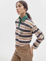 Remera tipo polo de manga larga con estampado de rayas horizontales en tonos beige, negro y naranja. Presenta cuello camisero verde, tapeta con botones y logo bordado en el pecho.