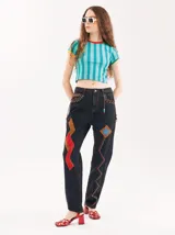 Pantalón de denim negro con corte mom-fit, tiro alto y diseño suelto. Presenta parches de gamuza en colores rojo, marrón y azul, detalles de tiento de cuero trenzado en los bolsillos frontales y adornos de canutillos.