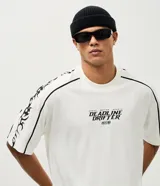 Remera de corte boxy y silueta amplia, color blanco con cuello redondo. Presenta estampa frontal con texto y gráficos tribales en las mangas.
