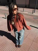 Campera tipo bomber color bordó, confeccionada en gamuza, con cierre frontal y una hilera de botones metálicos decorativos a lo largo de la solapa. Presenta mangas largas con puños holgados y un diseño de corte oversize.