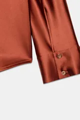 Camisa de manga larga confeccionada en tejido satinado, con cuello solapa, cierre frontal con botones y bajo asimétrico.
