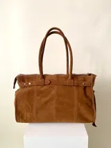 Bolso grande de cuero marrón con textura tipo cocodrilo, estructura rígida y asas para llevar al hombro. Tiene una tira con tachas y hebillas en cada extremo, también lleva lazos en los costados.