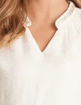 Blusa de satén color verde petróleo, con escote en V y cuello decorado con mostacillas bordadas. Presenta mangas largas con puños elásticos y ruedo también elastizado, brindando un ajuste cómodo y holgado.