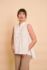 Blusa sin mangas de color blanco, con cuello mao y cierre frontal de botones. Presenta un diseño holgado y asimétrico, con la parte trasera más larga y aberturas laterales en el dobladillo.