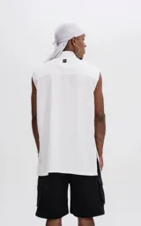 Camisa sin mangas de corte oversize, color blanco, con cuello clásico, cierre frontal con botones ocultos y bolsillo frontal con solapa y remaches metálicos.