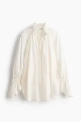 Camisa de lino y viscosa color blanco marfil, de diseño oversized, con cuello alto plisado y volados, mangas largas abullonadas con puños elásticos fruncidos y cordones ajustables en el escote.