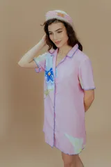 Vestido corto de lino y algodón color rosado, con cuello de solapa y manga corta. Presenta un diseño abotonado al frente con estampados de flores azules y saltamontes verdes, además de detalles de bordado a mano en contraste azul en los bordes.