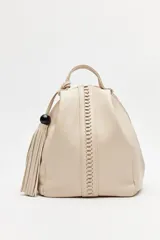 Mochila de símil cuero texturizado color beige, con asa superior, tiras regulables para la espalda y cierre principal metálico. Presenta un detalle trenzado vertical en el frente y un accesorio con flecos y cuenta negra en el lateral.
