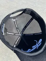 Gorra trucker gris con paneles frontales blancos y paneles traseros de rejilla negra. Presenta un logo bordado en el panel frontal y una cuerda decorativa en la base de la corona.