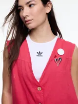 Chaleco sin mangas color coral, con escote en V, cierre frontal con botones de madera y bolsillos de parche. Incluye tres pins decorativos: un corazón plateado, un círculo plateado y un cuadrado rojo. Confeccionado en tejido liviano de lino y viscosa.