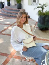 Cartera tipo sobre color crudo, confeccionada en cuero, con correa larga de cuero marrón y costuras decorativas en contraste.