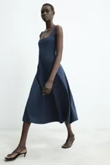 Vestido midi de color beige con escote cuadrado y tirantes anchos. Presenta un diseño entallado en el torso con costuras verticales marcadas y falda con caída evasé.