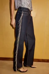 Jeans de tiro bajo y corte recto en denim color azul oscuro. Presenta cinco bolsillos, trabillas para cinturón y cierre frontal con cremallera y botón metálico. Destaca por la aplicación de flecos brillantes en los laterales de ambas piernas.