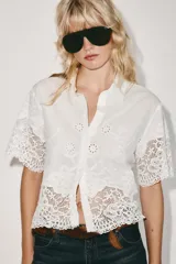 Camisa blanca de manga corta y cuello solapa, con detalles de bordados perforados y encaje en el borde inferior y las mangas. Cuenta con cierre frontal de botones.