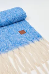 Bufanda rectangular de tejido de punto con textura jaspeada en color azul y flecos largos en color crudo en los extremos. Cuenta con una etiqueta de cuero sintético con logo en una de las esquinas.