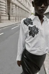 Camisa de cuello solapa y manga larga, con estampado de rayas verticales blancas y grises. Presenta dos aplicaciones de flores de encaje negro en la parte frontal. Cierre frontal con botones ocultos por solapa.