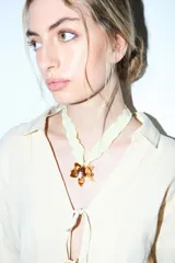 Collar con cinta de tela plisada color amarillo pálido, atado al cuello, con un dije colgante que simula una flor de hibisco en tonos marrones y dorados, con un detalle central perlado.