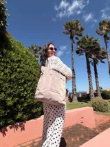 Bolso tipo tote bag de símil cuero color rosa pálido, con asas dobles y detalles de costura en color beige claro. Presenta un bolsillo frontal rectangular aplicado y herrajes metálicos.