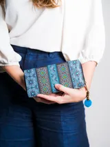 Billetera rectangular de tela bordada con motivos tribales en tonos de azul, marrón, verde y amarillo. Cierre con cremallera y tirador con cuentas de madera y pompón azul.