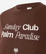 Remera de algodón color marrón oscuro, con cuello redondo y manga corta. Presenta un estampado frontal en blanco con el texto "Sunday Club Palm Paradise", una palmera y una frase motivacional.