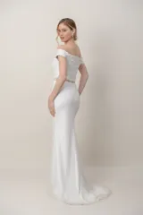 Vestido largo de novia de tul color blanco, con diseño de hombros descubiertos, mangas cortas caídas y corsé con apliques bordados a mano. Presenta una falda sirena con detalles florales bordados y cola.