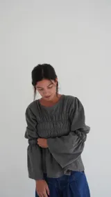 Camisa a cuadros azul y gris con cuello redondo y mangas largas abullonadas. Presenta detalle de fruncido en el frente y puños elásticos.