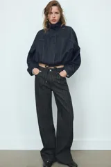 Cazadora de denim color azul oscuro, con cuello solapa, mangas largas tipo murciélago y bolsillos de vivo en el delantero. Presenta un bajo elástico ajustable con cordones y cierre frontal con cremallera y botones a presión ocultos.