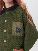 Campera tejido grueso y abrigado color verde militar, con forro de nylon interno, cuello estilo camisa en color negro y cierre frontal con botones. Cuenta con cuatro bolsillos delanteros con apliques en distintos tonos y texturas, y un parche bordado circular en el bolsillo superior izquierdo.