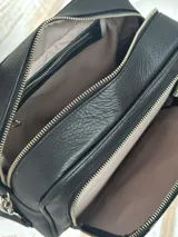 Bandolera negra de cuero con textura tipo cocodrilo en la parte superior. Tiene correa de tela negra con letras blancas y herrajes plateados.