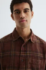 Camisa de franela de algodón suave con efecto lavado, color bordó con estampado a cuadros en tonos negros y amarillos. Presenta un corte relajado, cuello inglés, tapeta clásica de botones, un bolsillo superior abierto y canesú en la espalda. Tiene mangas largas con puños abotonados y un bajo ligeramente redondeado.