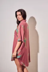 Vestido camisero corto de color rosado con estampado de aves abstractas en tonos verdes y azules. Cuenta con cuello camisero, cierre frontal con botones, mangas cortas y un bolsillo aplicado en el pecho.