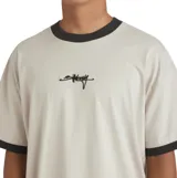 Remera Billabong modelo Script Ringer, de color beige con cuello redondo y ribetes en las mangas de color negro. Presenta un pequeño estampado gráfico central en el pecho.