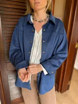Sobrecamisa de gamuza con cuello clásico, cierre frontal con botones metálicos y mangas largas con puños abotonados. Presenta un diseño estructurado con corte recto y largo a la cadera.