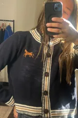 Saco de punto color bordó con detalles en color crema en el cuello, puños y borde frontal. Presenta un diseño de caballo bordado en el pecho y cierre frontal con botones oscuros.