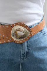 Cinto tipo faja de cuero color beige con tachas metálicas plateadas a lo largo de toda la pieza. Presenta una hebilla central grande de metal plateado con diseño grabado y una piedra natural incrustada en el centro.