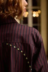 Camisa de algodón con diseño a rayas verticales en tonos oscuros, cuello clásico y cierre frontal con botones. Presenta detalles bordados en color amarillo a lo largo de las costuras laterales y en la parte superior delantera.