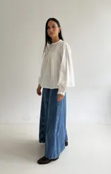 Blusa blanca de corte holgado con cuello redondo, mangas largas con puños elásticos y detalles de puntillas verticales en el frente.