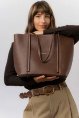 Bolso tipo tote de color marrón, confeccionado en material símil cuero texturado. Presenta un diseño estructurado con asas largas para llevar al hombro y el logo de la marca aplicado en la parte frontal.