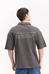 Polo de algodón color gris oscuro, de manga corta y corte oversize, con cuello solapa y cierre frontal corto con botones ocultos, y diseño texturizado con estampa pequeña en el pecho.