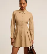 Vestido corto color beige, estilo chemise, confeccionado en tricolina. Tiene cuello inglés, mangas largas con puños abotonados, abotonadura frontal y falda con pliegues.