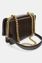Bolso mini bandolera de efecto acolchado, color marrón oscuro, con pespuntes verticales. Posee asa de cadena dorada que puede usarse como doble asa de hombro. Cierre mediante solapa y botón imantado.